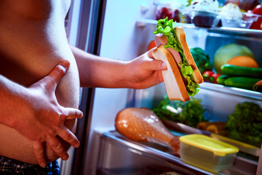 Hungry man holding a sandwich in his hands and standing next to the open fridge.