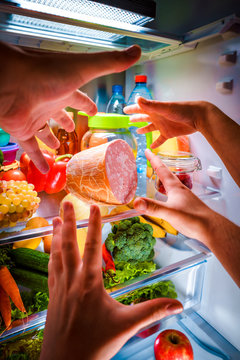Human hands reaching for food at night in the open refrigerator