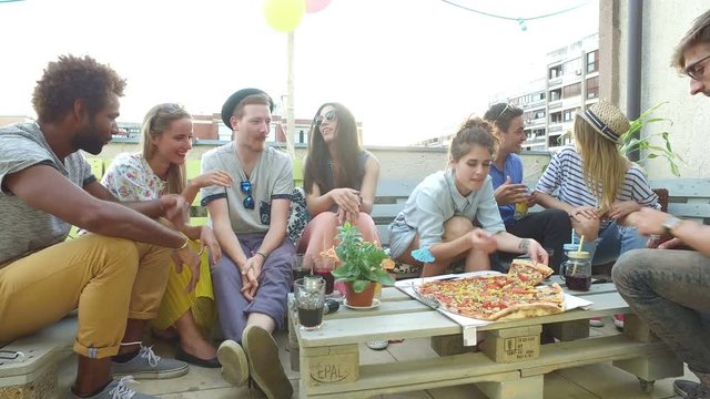 Attractive young people sitting around the table on the rooftop terrace, preparing to eat pizza