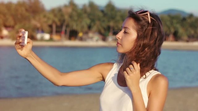 Happy young woman making selfie on seaside beach, taking picture on smartphone