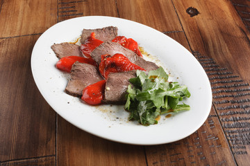 Baked roast beef with rucola and red bell pepper in a white oval plate. Brown wooden background.