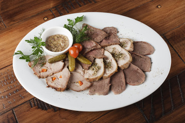 Baked roast beef, chicken rolls, tongue and pork meat with vegetables in a white oval plate. Cold appetizer with sauce. Wooden background. 