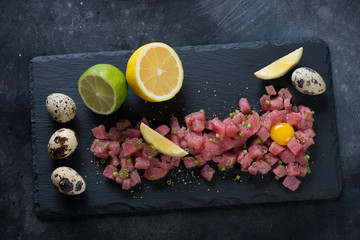 Fototapeta na wymiar Stone slate tray with fresh tuna tartare, quail eggs, lemon and lime, above view