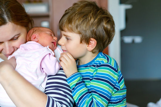 Little kid boy kissing newborn sister. Mother holding baby on arm.
