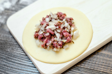 Circle of raw dough with meat and onions, on a wooden board, and flour