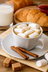 Cup of coffee with marshmallow, cinnamon sticks, brown sugar and croissant on wooden table.