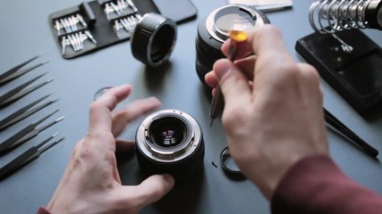 Hands with screwdriver repair photo camera on grey table, close up. Unrecognizable professional technician with tool carefully remove two small screws of lens on lit workplace with instruments.