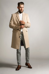 Elegant bearded man in a suit and in a beige coat, standing on a gray background