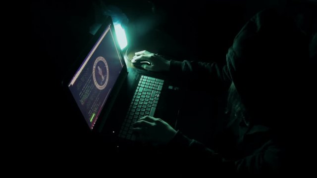 Hacker in hood cracking code using laptop and computers from his dark hacker room