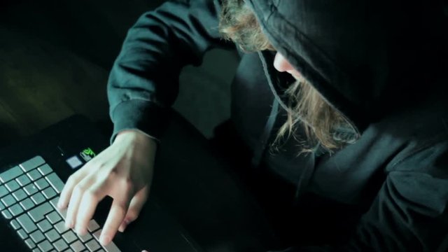 Hacker in hood cracking code using laptop and computers from his dark hacker room