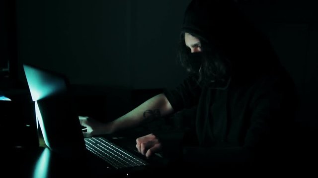 Hacker in hood cracking code using laptop and computers from his dark hacker room