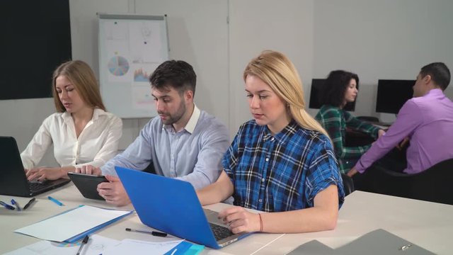 Creative young business team working in the room together. Two attractive caucasian blonde looking in tablet with happy smile. Group of people using modern device typing on laptop. On the background