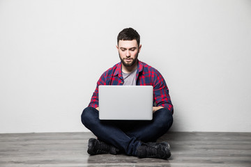 Young man in interior with white wall sitting on wooden floor with laptop