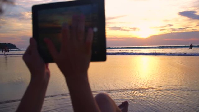 Young woman taking pictures of sunset over ocean using tabler computer.