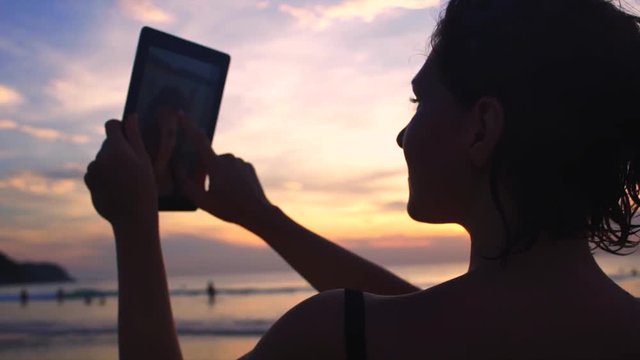 Young woman taking selfie with tablet at sunset by the ocean