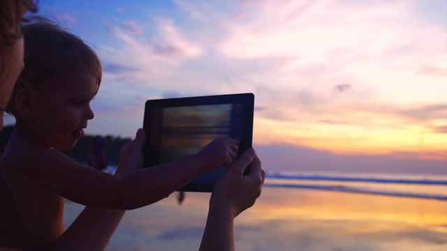 Young woman with her baby taking pictures of sunset over ocean using tablet computer.