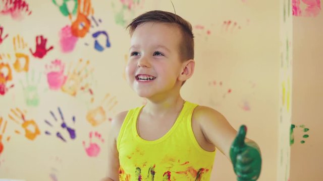 Close up veiw of cute little boy with painted hands after leaving his colorful handprints on the wall and showing his thumb up. Young happy family. Mother and child concept