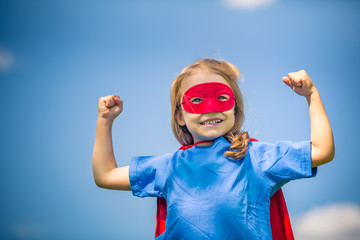 Funny little girl playing power super hero over blue sky background. Superhero concept.