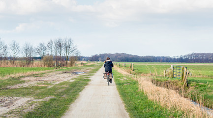 Woman with black jacket bikes on a dirt road in a flat agricultural landscape in the Netherlands. Spring has just begun.