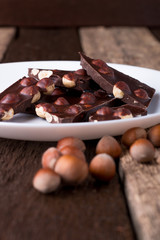 Chocolate pieces with hazelnut nuts on white plate wooden background.