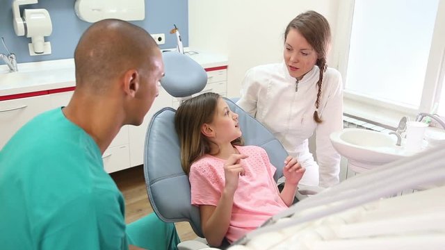 Lovely little girl sitting in the dental chair, talking with dentist and assistant