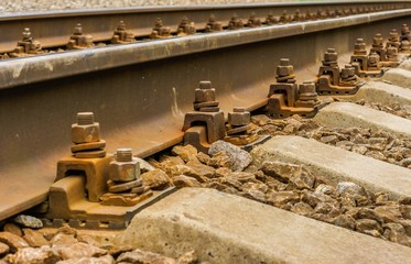 Fototapeta na wymiar Background picture: steel rails which run diagonally from lower left corner to the upper right corner close-up visible rusty fasteners, nuts, bolts, stones, concrete sleepers