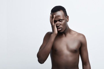 Fototapeta premium Przestraszony zmartwiony Afroamerykanin o ciemnej karnacji pozuje na szarej ścianie studia, patrząc prosto w kamerę przez dłoń na twarzy. Pojęcie negatywnych emocji.