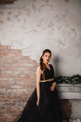 Fototapeta na wymiar beautiful brunette girl in long black dress makeup red lips and manicure is standing near the fireplace looking down