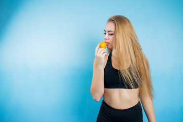 copyspase beautiful girl in sportswear short shorts and top black on a blue background holding a tangerine and is about to eat looks at the fruit