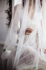 beautiful girl in white transparent boudoir gown of tulle and lace closed with a veil sitting on the windowsill near the window