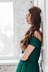 beautiful brunette girl with long hair standing in green dress near the white wall by the window looking to the side