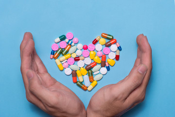 Colored pills in heart shape covered with hands on blue background.