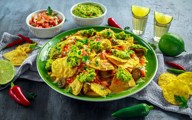 Nachos with cheese, jalapeno peppers, red onion, parsley, tomato, salsa, guacamole sauce and tequila on green plate.