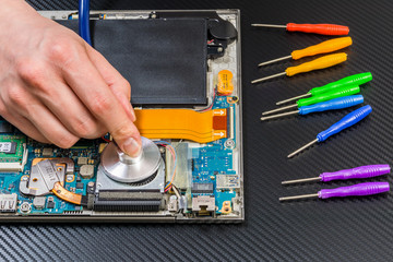 Fototapeta na wymiar person hands diagnostic laptop mainboard with stethoscope on carbon background with rainbow collection of screwdrivers