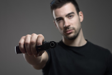 Dangerous criminal holding tilted gun threatening aiming at camera. Atmospheric contrasty portrait over gray studio background.