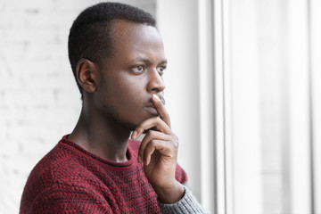 Fototapeta na wymiar Pensive worried African American art director dressed casually keeping finger on lips while thinking over something important, trying to find creative solutions for current issues, standing at window