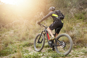 Back view of male rider in protective gear riding e-bike in countryside, getting over obstacles on his way while cycling among trees, exercising his freeride skills, sun shining brightly in background