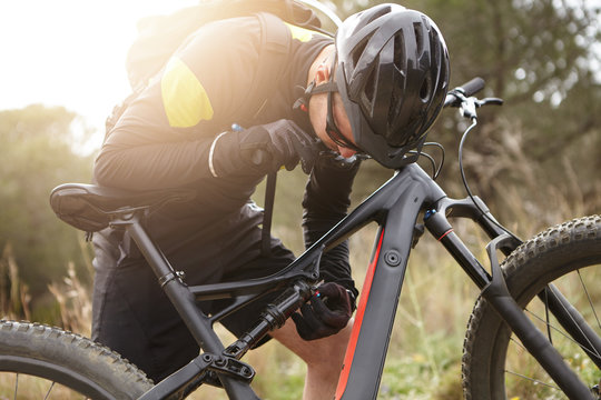 Candid view of male cyclist in sportswear, protective helmet and glasses fixing his motor-powered electrobike that he broke during freeride in woods. Rider repairing his booster bicycle outdoors