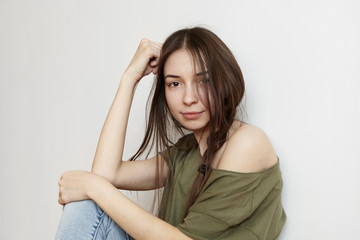 People, youth and happiness. Beautiful girl of tender age sitting on floor, spending leisure time at home after college, dressed in jeans and oversize army green top with one shoulder showing up