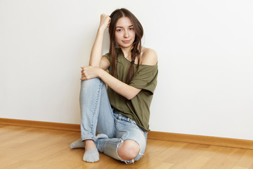 Indoor shot of beautiful teenage girl with messy hair wearing ragged jeans and stylish oversize top sitting on floor in her white bedroom, spending weekend at home and waiting for friends to come