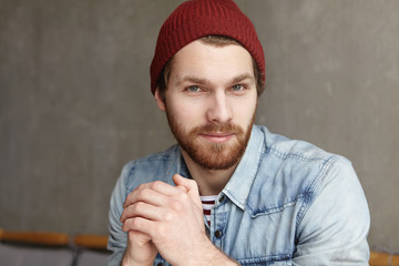 Handsome young Caucasian man with thick beard looking at camera with flirting smile, keeping hands clasped. Attractive guy wearing stylish clothes, sitting isolated against concrete wall background