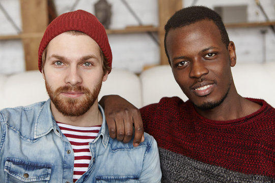 People, love and homosexuality. Samesex male couple spending nice time together at cafe: Afro-American man holding hand on his Caucasian partner's shoulder, both looking and smiling at camera