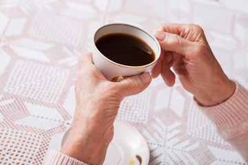An elderly woman drinks tea at home. Senior woman holding cup of tea in their hands at table closeup. Horizontally top view