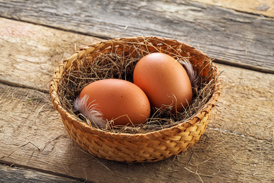 Two fresh farm eggs in a basket with hey and feather on a wooden table. Close-up shot. Top view.