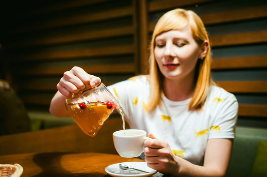 Young blonde woman wearing white T-shirt with print, girl pours a fruit drink from a glass jug, into a white cup, sitting at a table in a cafe, background stylized Wood texture lit up with warm light