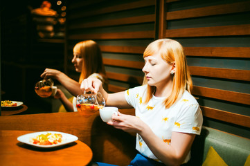 Young blonde woman wearing white T-shirt with print, girl pours a fruit drink from a glass jug, into a white cup, sitting at a table in a cafe, background stylized Wood texture lit up with warm light