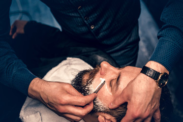 Barber shaving bearded male with a sharp razor.