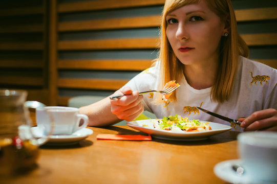 Young blonde woman wearing white T-shirt with print, girl Eats salad at lunch time, sitting at a table in a cafe, background stylized Wood texture lit up with warm light