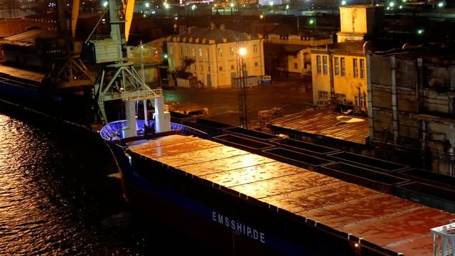 top view of empty long barge and ship on the dark water in the seaport with buildings and constructions under the lights during night