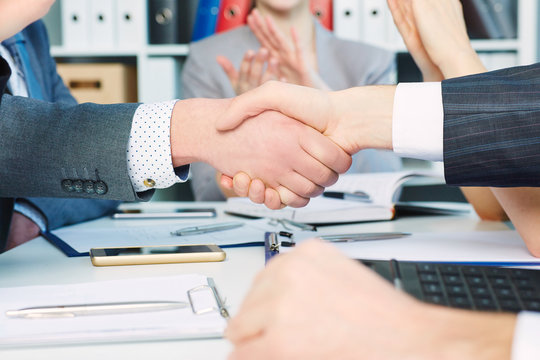 Close up businessmen handshake on team meeting with clapping group of people blured in background at modern startup business office interior.